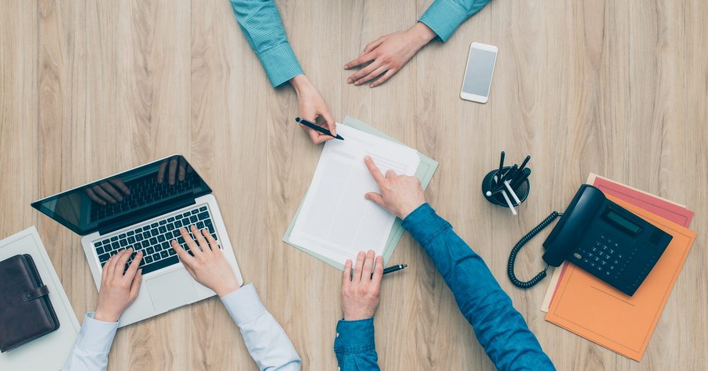 Kolme henkilöä kokouksessa pöydän ympärillä tietokoneen, puhelimen ja asiakirjojen kanssa