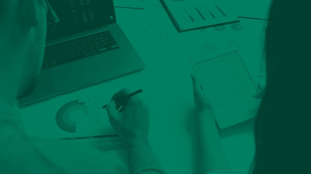 Tietokone pöydällä ja henkilö kirjoittaa muistiinpanoja